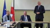 Zdjęcie sali. Trójka mężczyzn za stołem. Dwóch po lewej siedzi, mężczyzna po prawej stoi. W tle są flagi Polski oraz Unii Europejskiej.