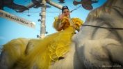Na środku zdjęcia kobieta w żółtej sukience. Włosy ma związane w kucyk. Na twarzy makijaż z cekinami. Siedzi na białym koniu. 