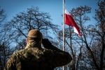 Sylwetka salutującego żołnierza tyłem. W dalszym planie powiewa flaga Polski na maszcie. 