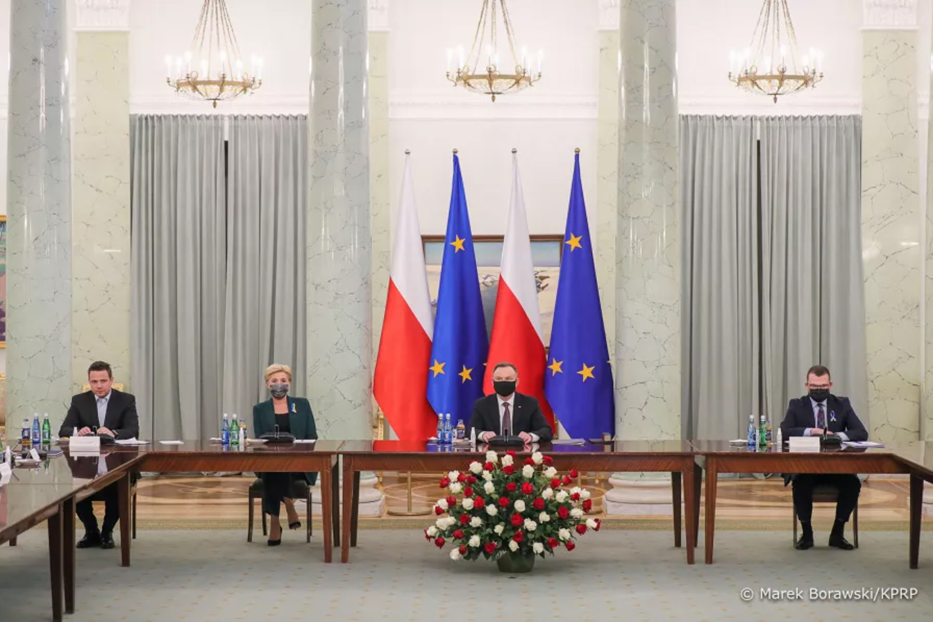 Na zdjęciu widać cztery osoby siedzące podczas obrad. Siedzący siedzą na tle czterech flag, dwóch Polskich oraz dwóch Unii Europejskiej